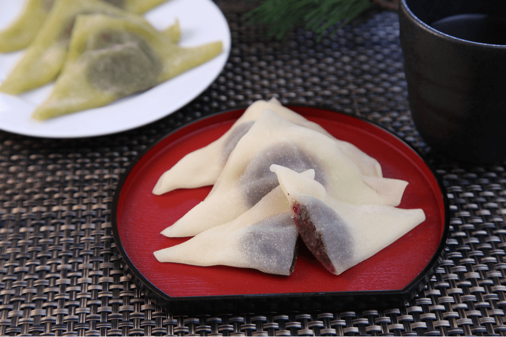 Six pieces of yatsuhashi, one of the Kyoto types of mochi, with plain and green tea flavor mochi folded over red bean paste on plates on a black placemat.