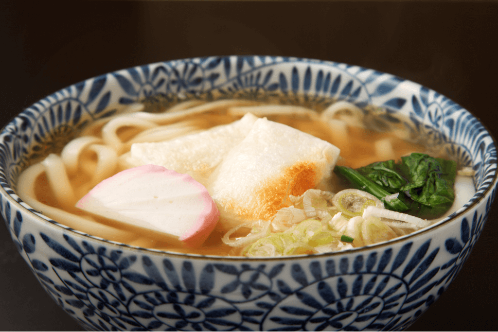A bowl of Chikara Udon, a udon noodle soup with pieces of grilled mochi, Japanese fish cakes, onions, and spinach, in a blue and white bowl.