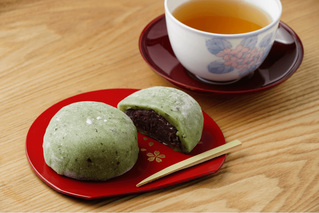 Two pieces of kusa mochi, another one of the stuffed mochi dish with a green instead of white mochi, on a plate, with one cut in half, next to a cup of tea