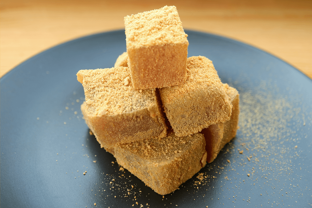Cubes of warabi mochi, a type of mochi made with fern starch and covered in soybean dust, stacked on top of each other on a black plate.