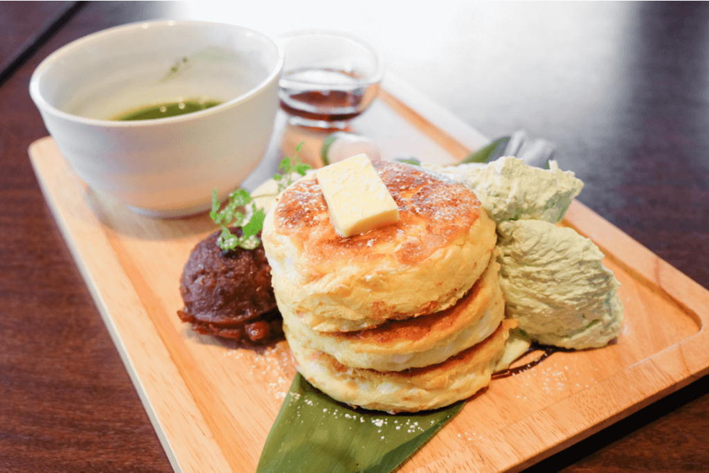 Japanese fluffy pancakes made with mochi sit on a wooden plate alongside ice cream, red bean paste, and a bowl of green tea on a cafe table.