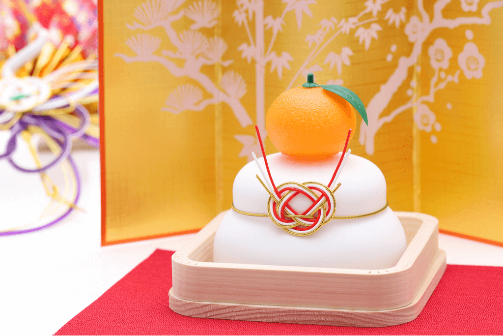 A replica of Kagami mochi sits in a wooden box with a tangerine and a string on top in front of a gold folding screen.