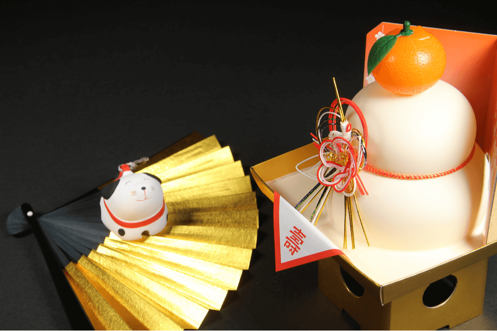 Kagami mochi sits on a golden pedestal with an intricate ribbon on white, red, and gold, in front of  a white and black fan with a dog figure sitting on the fan.