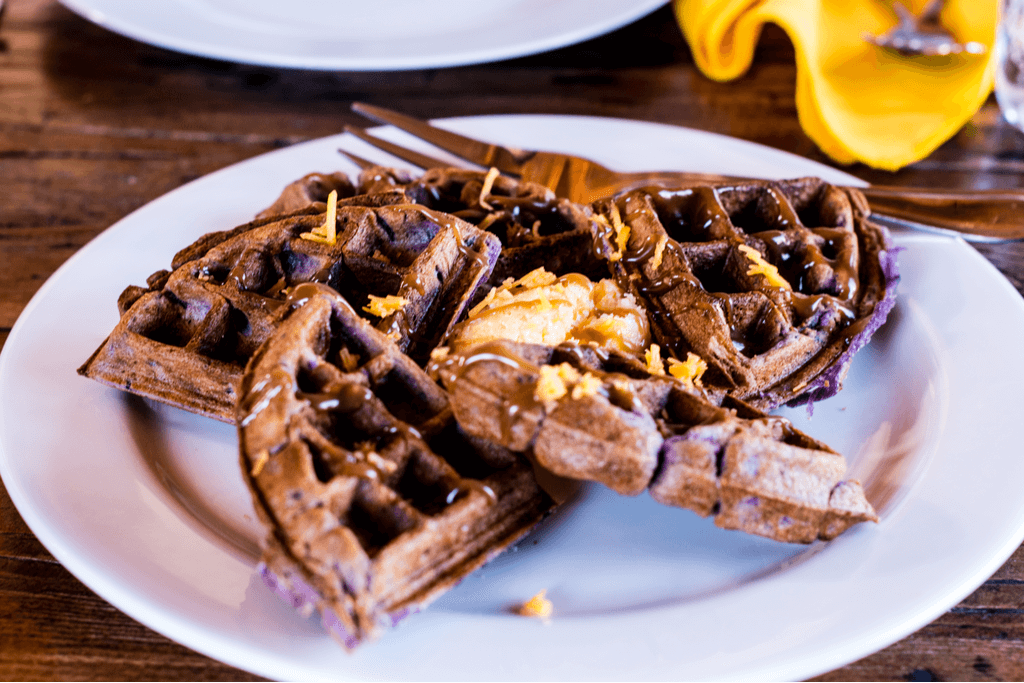 Brown waffle sections with hints of purple, made with a mochi and ube batter, sits on a plate next to a fork with a mochi cake in the middle.