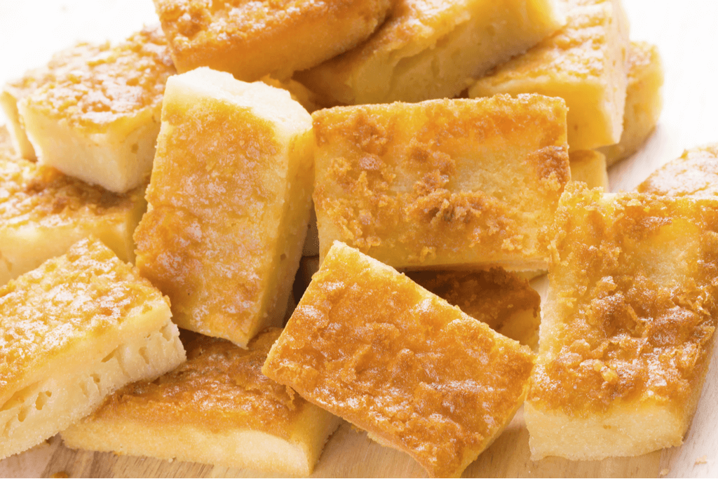 A pile of Hawaiian mochi desserts called mochi cake or butter mochi sits on a light brown table with many of the cakes showing a golden-brown crust.