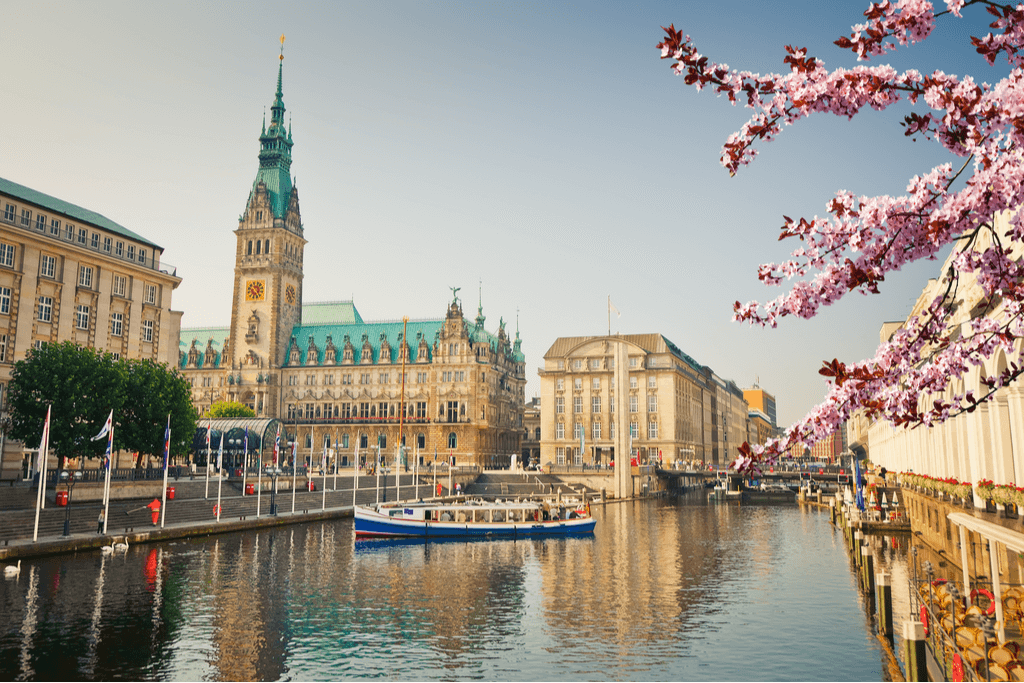 A photo of a boat on the river running through Hamburg, Germany, one of the best cities for Japanese food in the world. with buildings in the background and a tree of blossoms on one side.