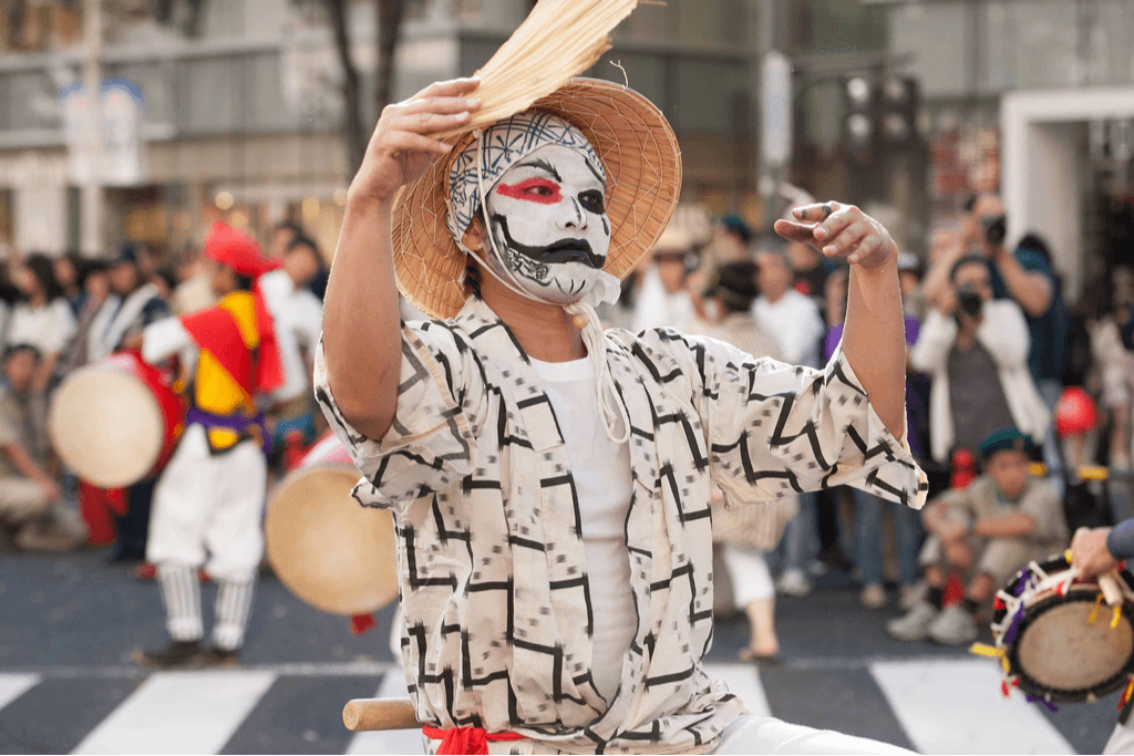 A man in a traditional garb with a fan and a painted face dances at the Gion Matsuri on the street.