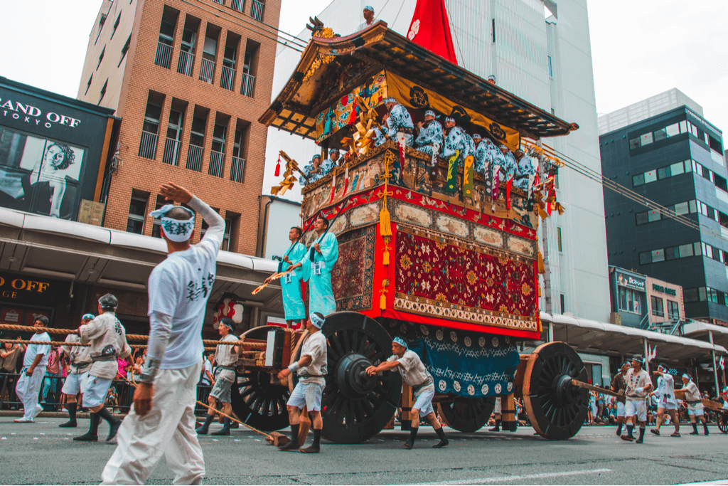 Men pull a giant Gion Matsuri float with even more people riding it a man directing them with a fan.