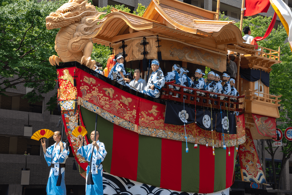 Two men in kimono direct a giant Gion Festival float with a boat-like shape as different men sit on the float as well.
