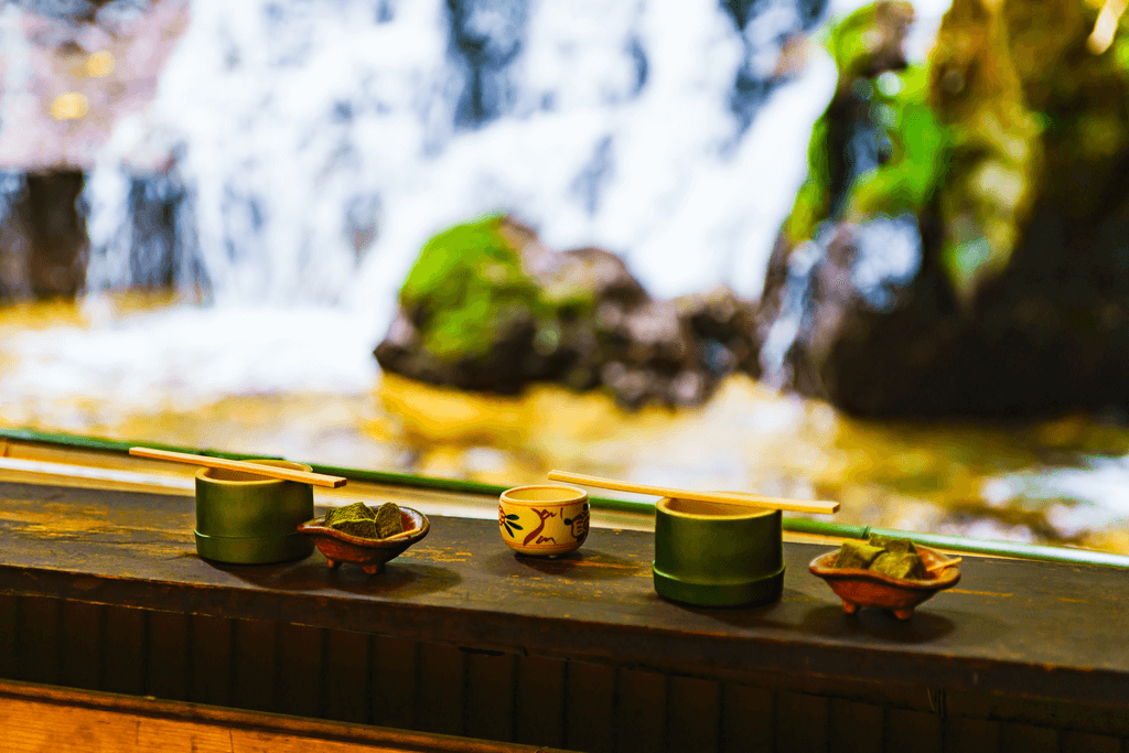A photo of one of the counters at Hirobun, a nagashi somen restaurant, with a waterfall in the background and bowls of tsuyu, cups of tea, and pieces of mochi in a bowl on the counter.