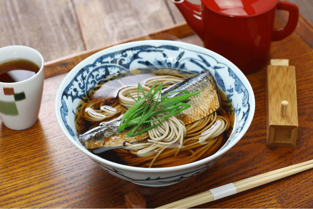 A bowl of Nishin Soba sits on a tray next to chopsticks and tea, with noodles sitting in a darker broth and a piece of mackerel laid over it with a garnish of green onion.
