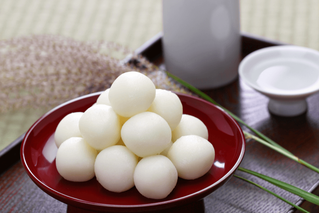 Tsukimi dango, white balls of mochi, sit in a pyramid shape on a red plate next a bottle of Japanese sake and a sake cup.