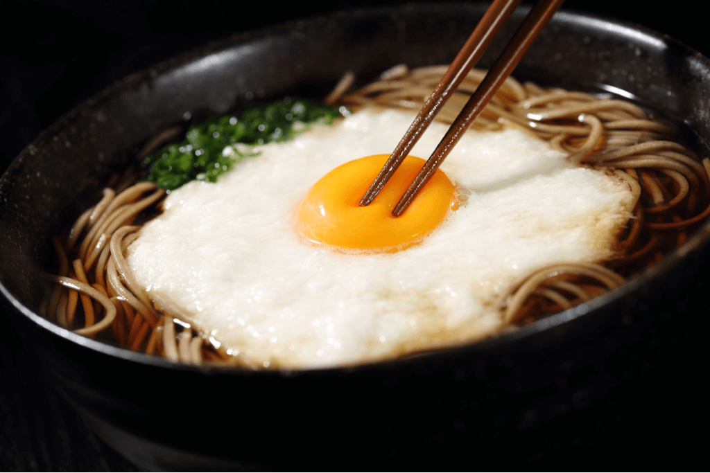 A black bowl of tsukimi soba, a soba noodle dish with an egg on top, with chopsticks on top pressing into the egg's yolk.