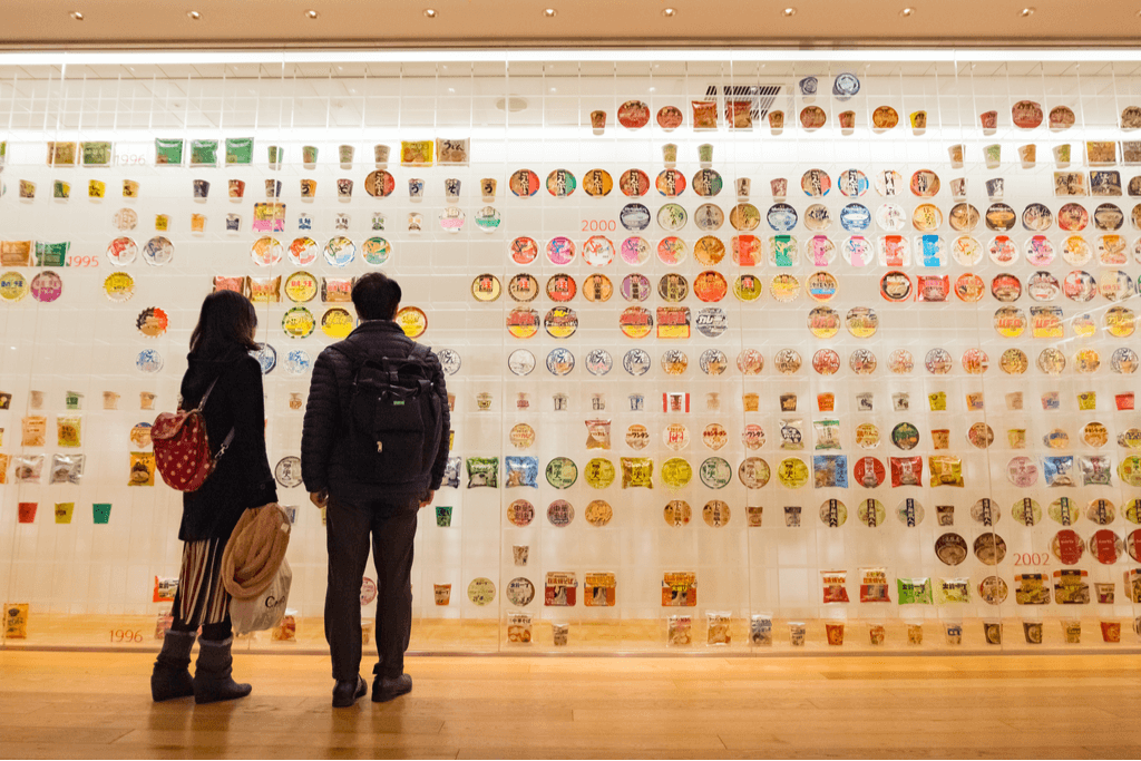 A woman and man stand in front of a wall of cup noodle at the Yokohama Cup Noodle Museum, one of two Yokohama museums about ramen