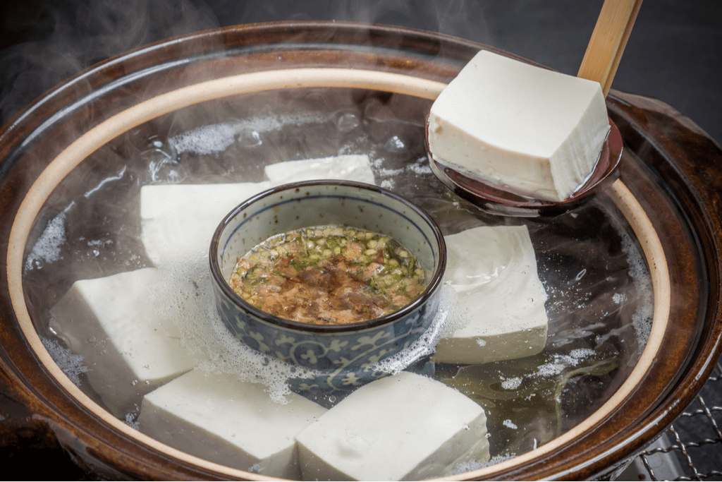 A hot pot filled with a boiling broth with blocks of tofu, one of the most famous Kyoto foods, with a ladle fishing out one of the blocks, and a cup of some kind of soup in the middle.