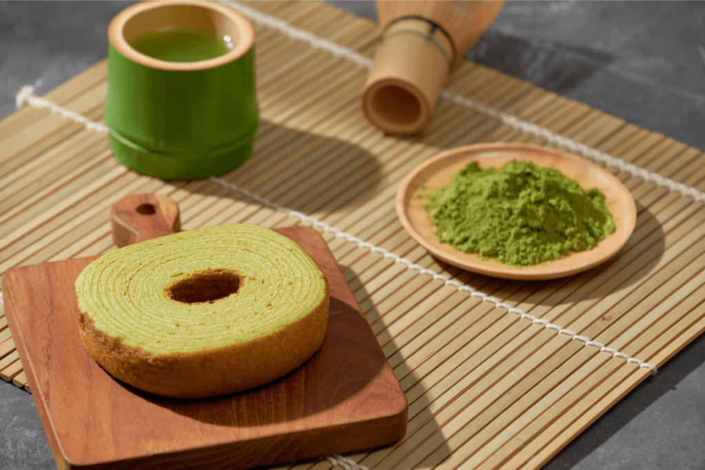 A photograph of a plate of Kyo baum which is matcha green tea-flavored baumkuchen (tree cake) along with some matcha powder and a wooden whisk on the side.