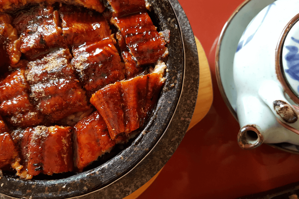 A close up photograph of grilled eel smothered in sweet soy sauce. A small teapot is next to it.
