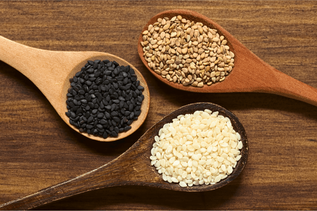 Three brown spoons, one with black sesame seeds, one unhulled white sesame seeds, and white sesame seeds