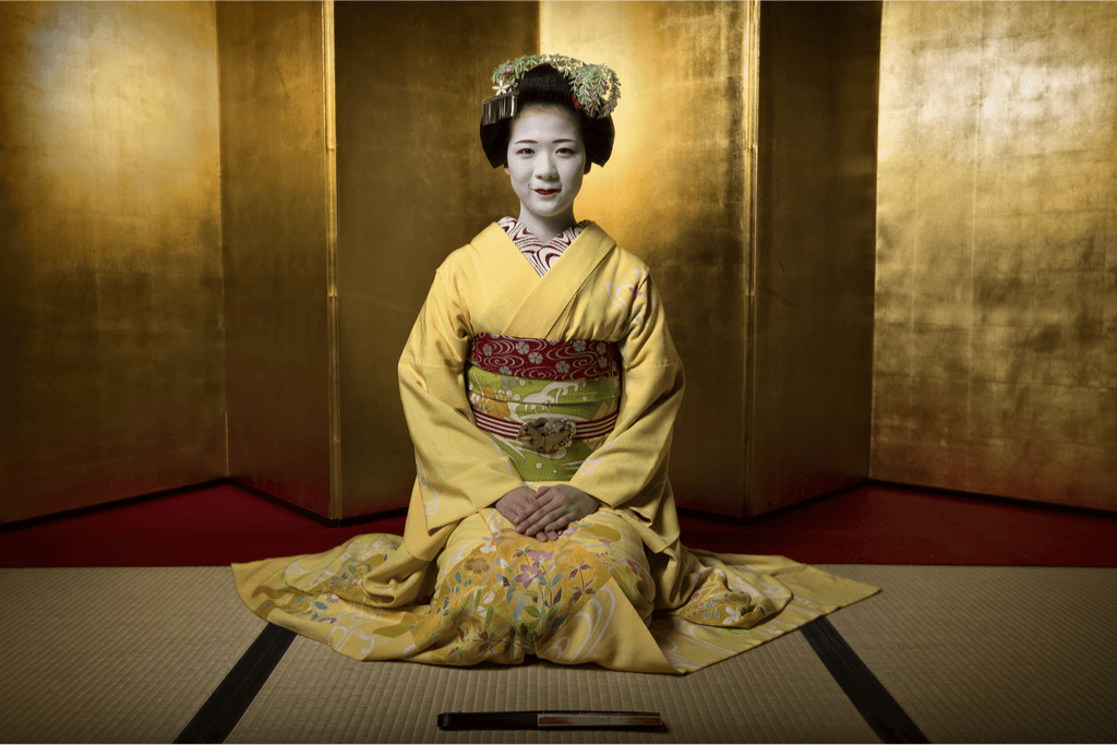 A Kyoto geisha sits in the center of the tatami room, dressed in an elegant yellow kimono and white makeup.