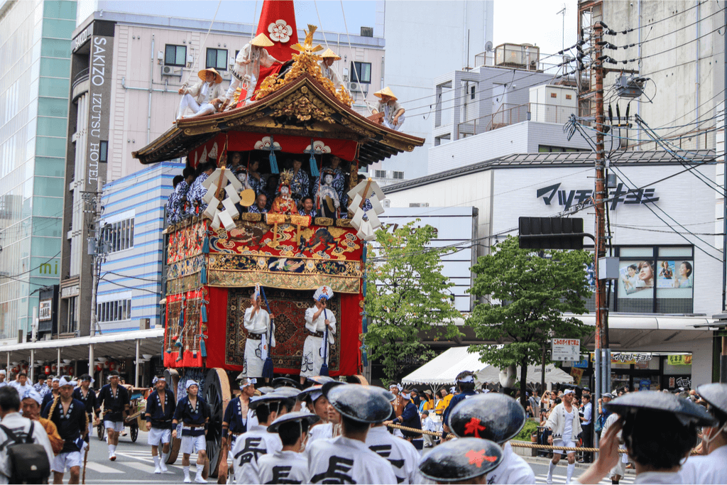 A group of shrine pullers dressed in all white, pulling an ornate shrine in the street. The ornate shrine is tall and orange.
