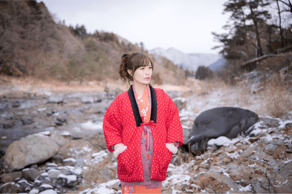 A woman standing in a river wearing a red, padded hanten winter jacket.