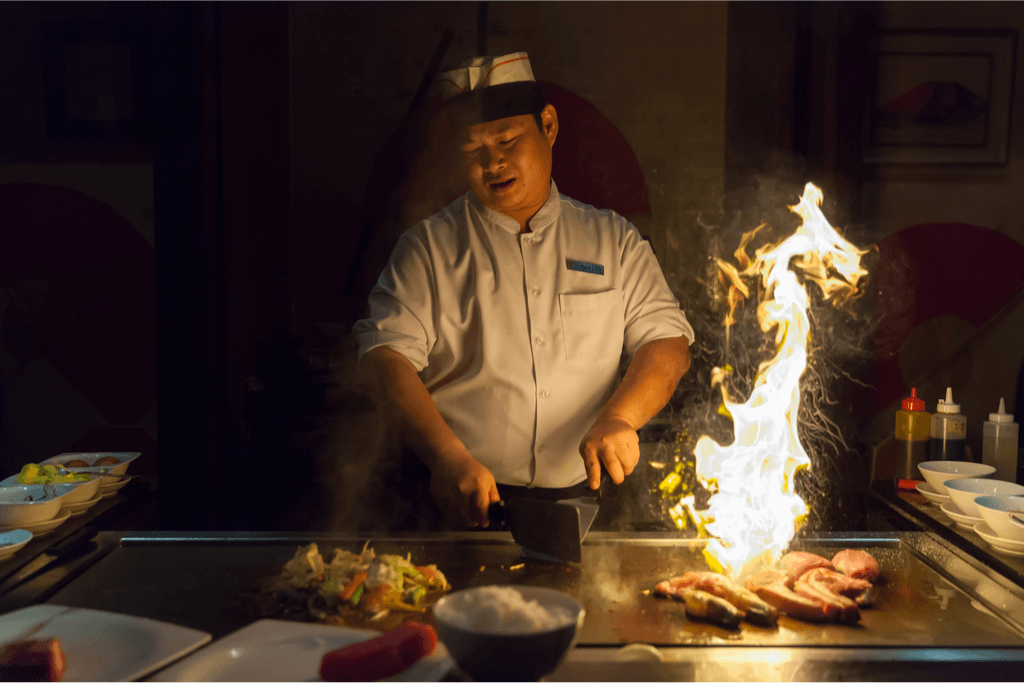 An chef cooking American-style hibachi food on a teppanyaki grill. There are a lot of flames.