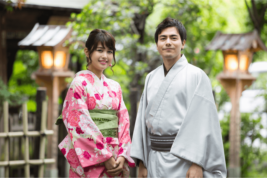 A man and a woman standing side by side wearing a pink/white and grey yukata respectively, a form of tradtional Japanese clothing.