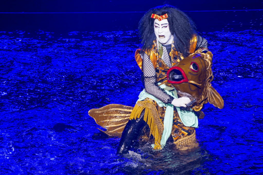 A Kabuki actor costumed in a long black wig, dramatic white makeup, is holding a lifesize goldfish prop, contrasting a against a deep watery blue stage.