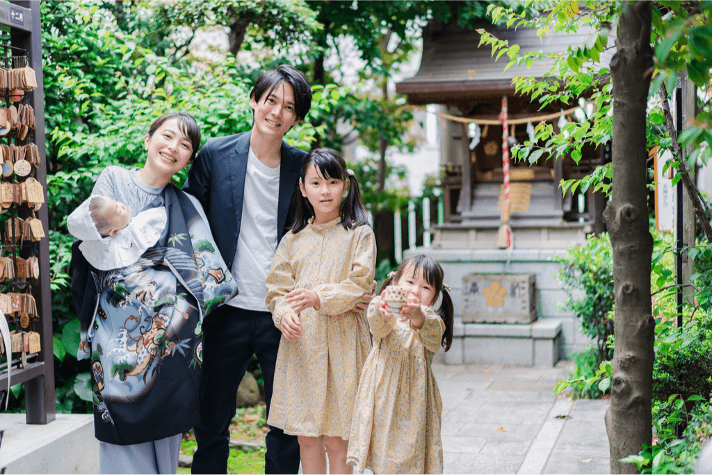 A family of five (mother, father, two youn daughters and a baby) are posing for a snapshot outside of a Shinto shrine.