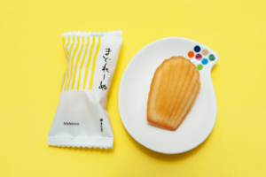 A madeline (long, shell-shaped pastry) on a plate, next to its original packaging, against a yellow background.