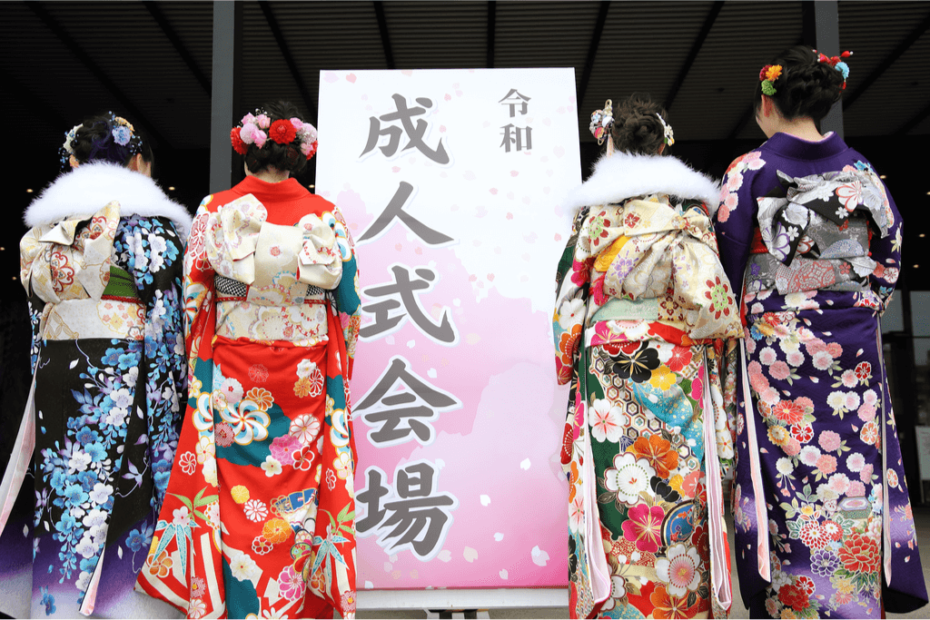 Four young women dressed in elaborate furisode kimonos with fur collars stand outside a sign for a Coming-of-Age Ceremony. It is a crucial part of Shinto in Japan.