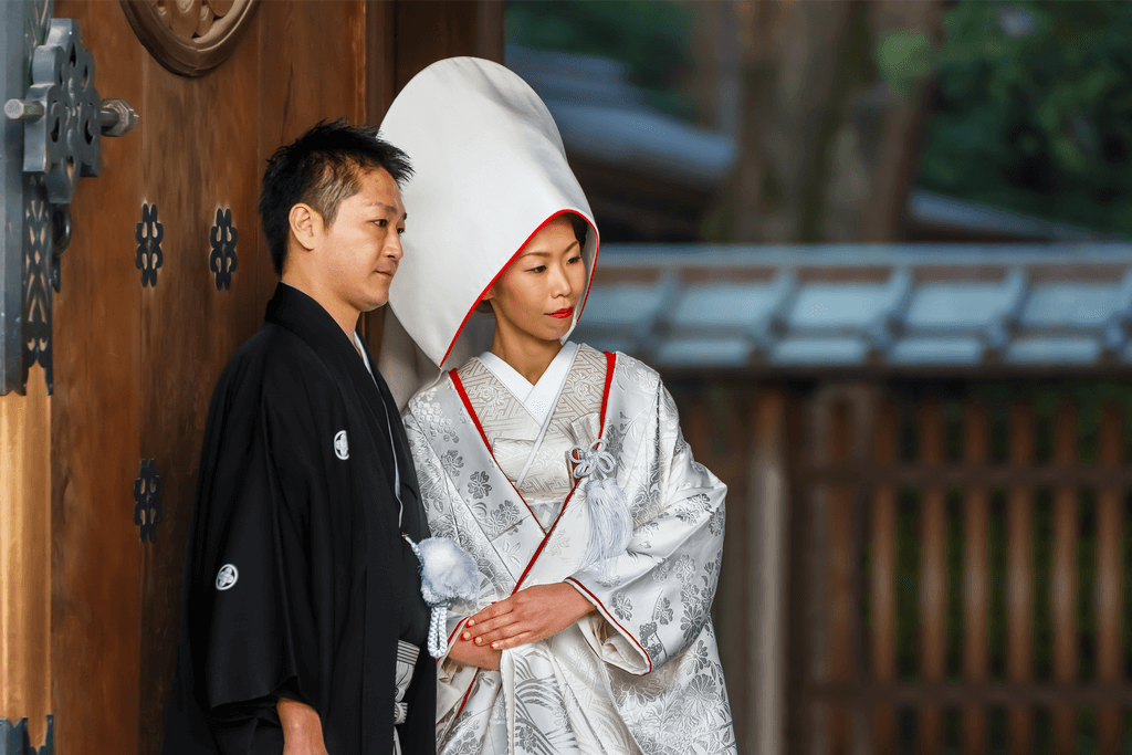 A bride and groom at a traditional Shinto wedding. The groom is wearing a black kimono and grey hakama, while the bride is wearing a white uchikake jacket and a white shiromuku kimono.muku