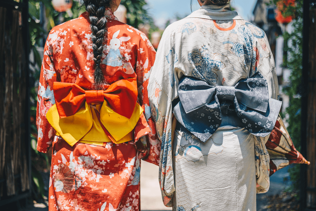 Two women wearing yukata with their backs facing the camera. The one on the left is wearing red with a yellow obi sash, while the one on the right is wearing a cream yukata with a grey obi.