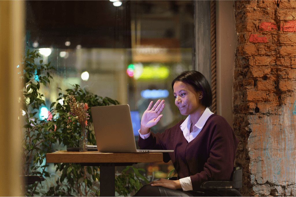 A woman sitting in a cafe in the evening, greeting someone on her laptop.