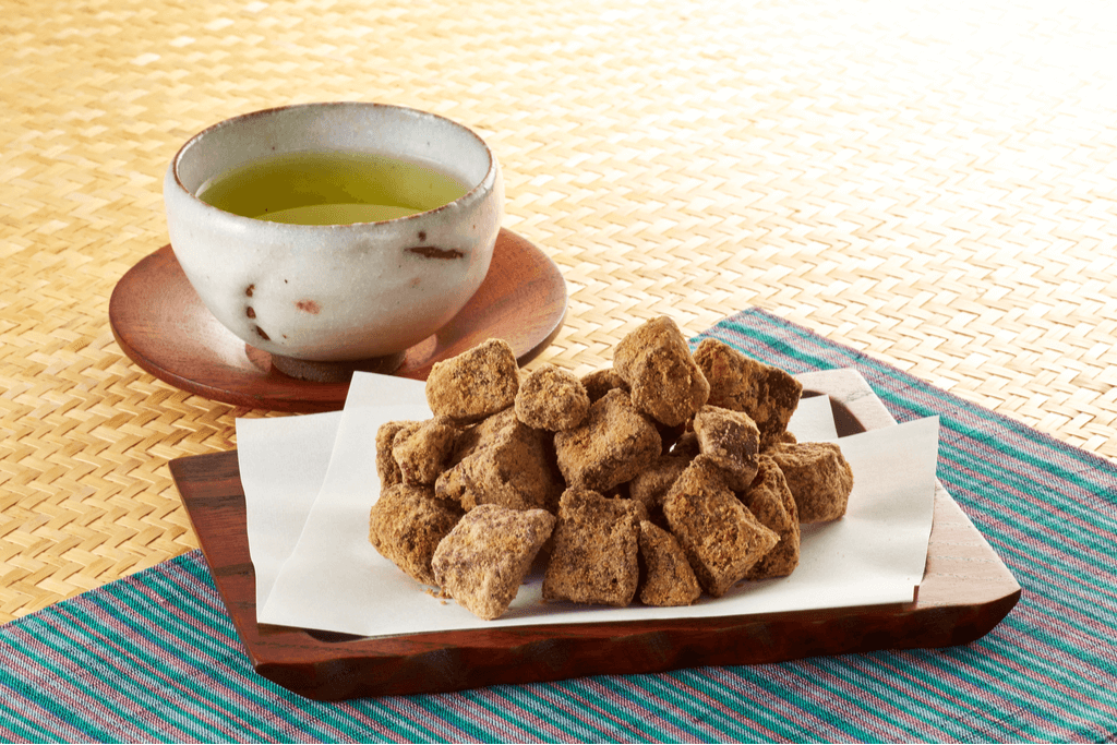 A plate of kokuto (dark brown sugar) cubes on a plate, with a cup of green tea slightly behind it. Kokuto is one of Okinawa, Japan's most treasured exports.