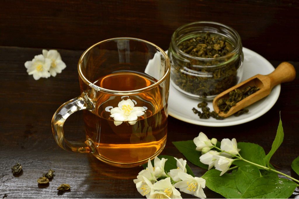 A host of jasmine tea, jasmine flowers and a scoop of jasmine tea leaves in a glass, jar and scoop, respectively. Jasmine flowers are light yellow and jasmine tea is light brown.