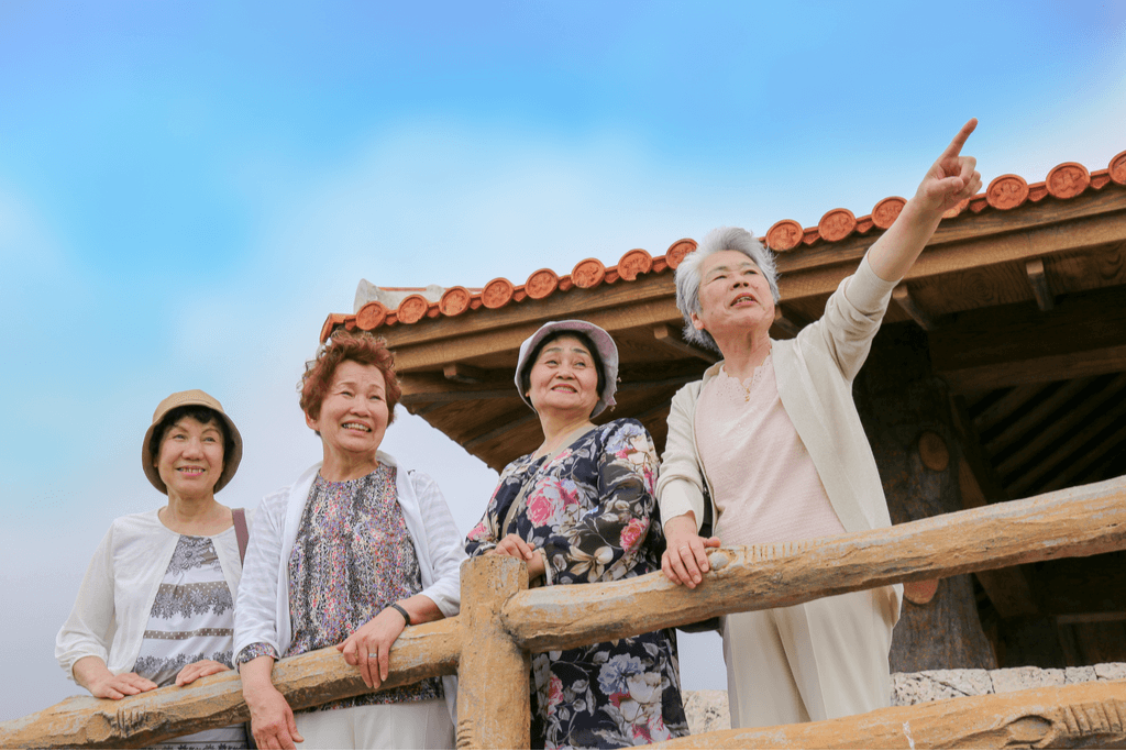 Four elder Japanese women happily standing outside a house, near a wooden railing. One of them is pointing to something ahead of her, offscreen. Community and purpose is a mainstay in Okinawa, Japan.