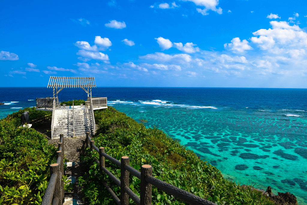 A daytime shot of a lush green hill and a gray pillar near a clear, blue beach.