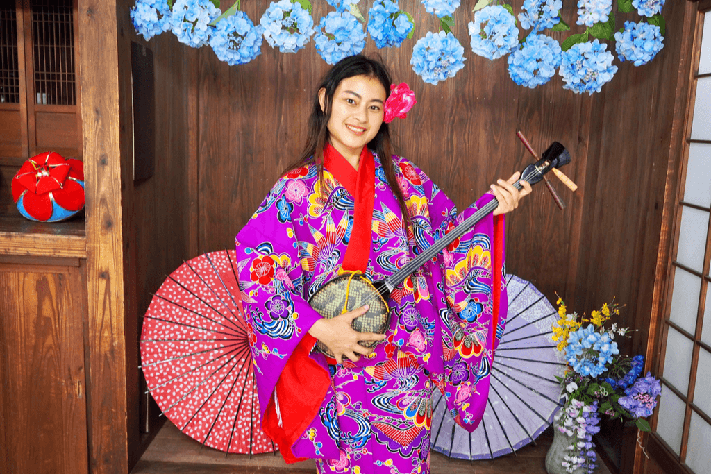 A woman dressed in a colorful ryusou (traditional Okinawan dress that resembles a robe), while playing a sanshin (Okinawan banjo). 