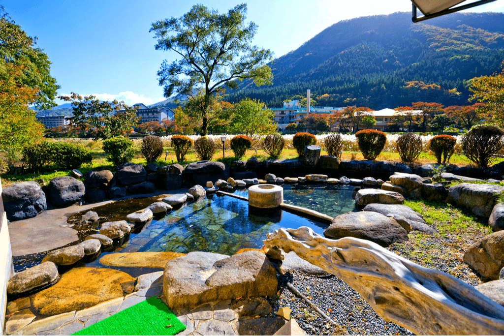 A daytime shot of Shiobara Onsen. The hills are green, the stones are grey and the water is a dark blue.