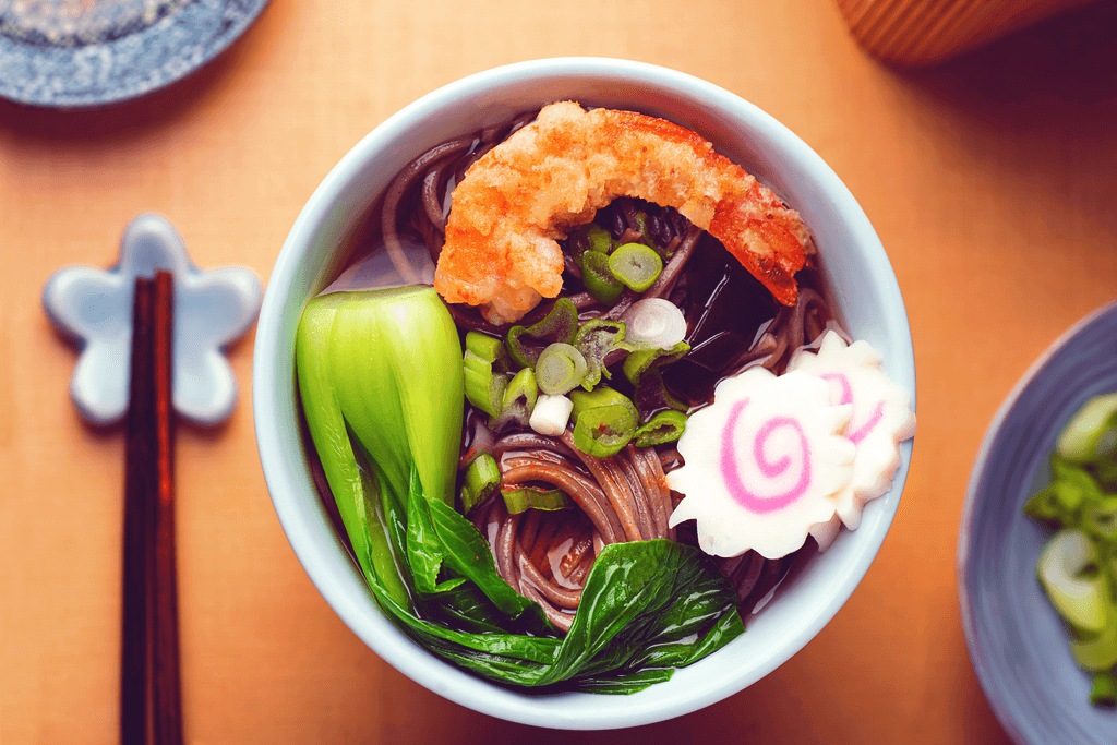 A bowl of soba with tempura shrimp, naruto fish cakes, shrimp and a whole stalk of spinach.