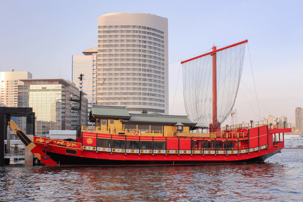 A picture of a yakatabune pleasure boat with a skyscraper in the background. The boat is red.