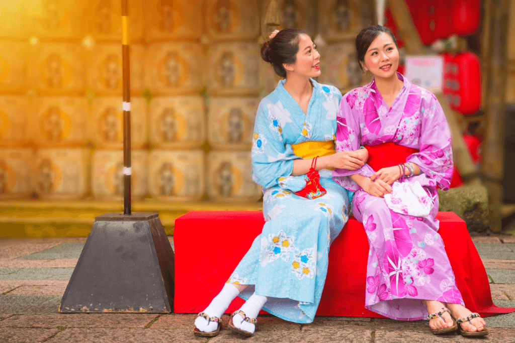 Two women in blue and pink yukata respectively, sitting on a red bench.