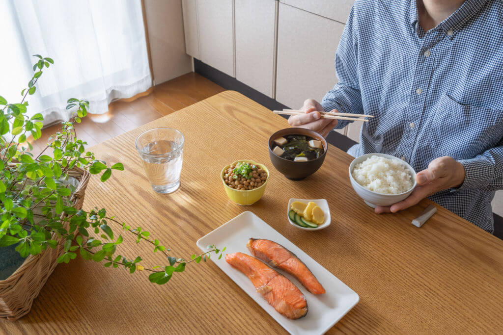 A man eating a salty Japanese breakfast. He eats white rice and miso soup. Side dishes include grilled salmon, natto, and pickles.