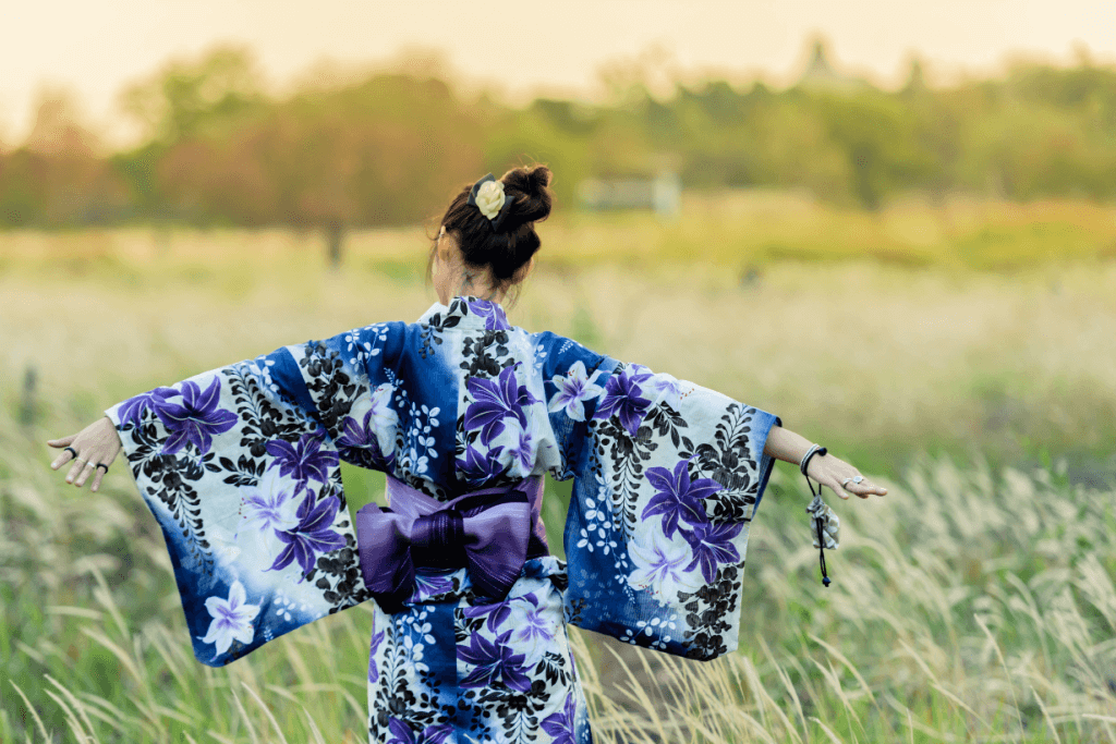A rear view of a woman wearing a blue yukata in a field, with her arms outstretched.
