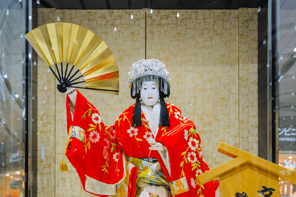 A feminine bunraku puppet in a red kimono holding a golden fan.