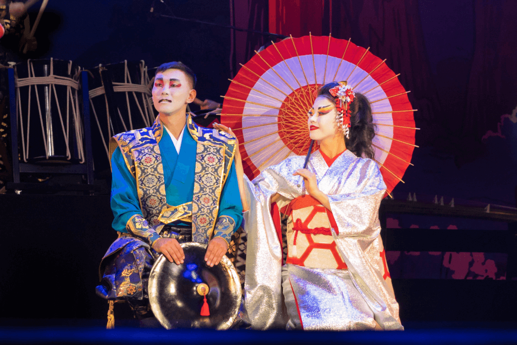 Two kabuki actors in a scene: one is in a masculine role wearing a blue kimono, while the other is in a feminine role wearing a kimono while holding a parasol. Kabuki is more lively than noh theater.