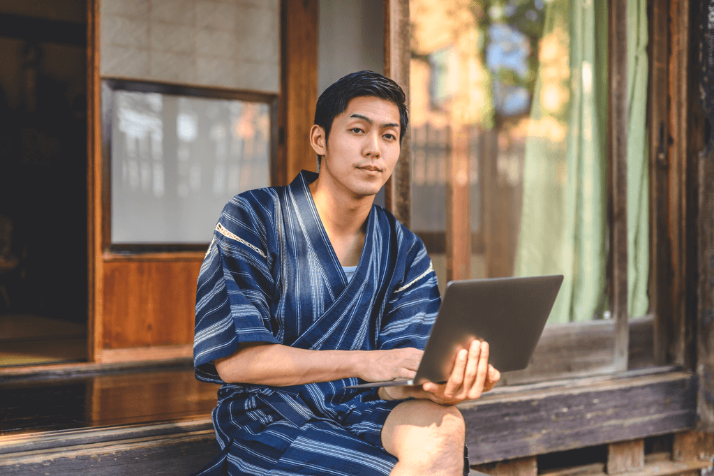 A man wearing jinbei clothing on the porch in the summer, while holding a laptop.