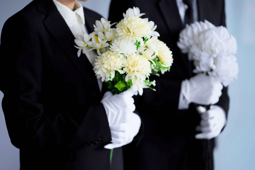 A man and woman using black suits at a funeral wearing white gloves and holding bouquets of white chrysanthemum.