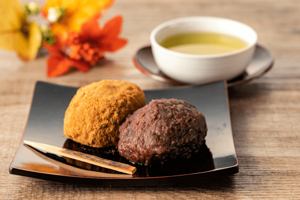 Two pieces of ohagi (sweet paste coated rice balls) on a plate next to autumn leaves.